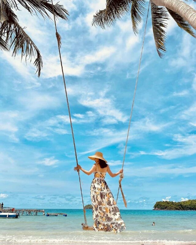 Tạo dáng đứng trên xích đu với hai tay nắm hai dây của xích đu, góc chụp sau lưng với mặt quay nghiêng hướng ống kính máy ảnh, xích đu nhẹ đưa làm váy tung bay