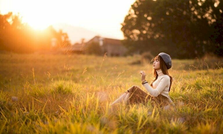 tạo dáng ngồi nghiêng trên cánh đồng cỏ, hai chân co nhẹ, một tay chống để người hơi ngả ra sau, một tay cầm một nhánh cỏ đưa lên mặt, mặt hơi hướng cằm lên trên với mắt nhắm nhẹ