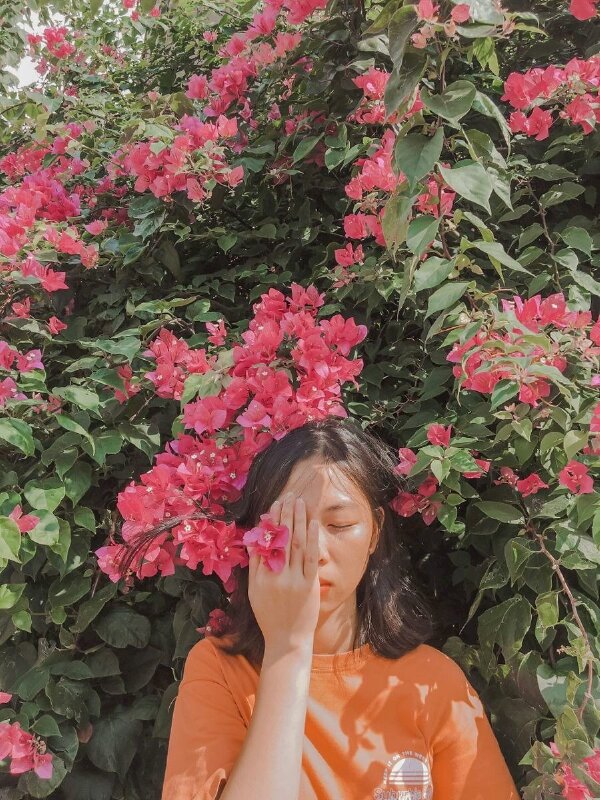 Tạo dáng đứng thẳng hướng về phía ống kính, dùng một tay có kẹp nhành hoa giấy che mặt, mắt khẽ nhắm